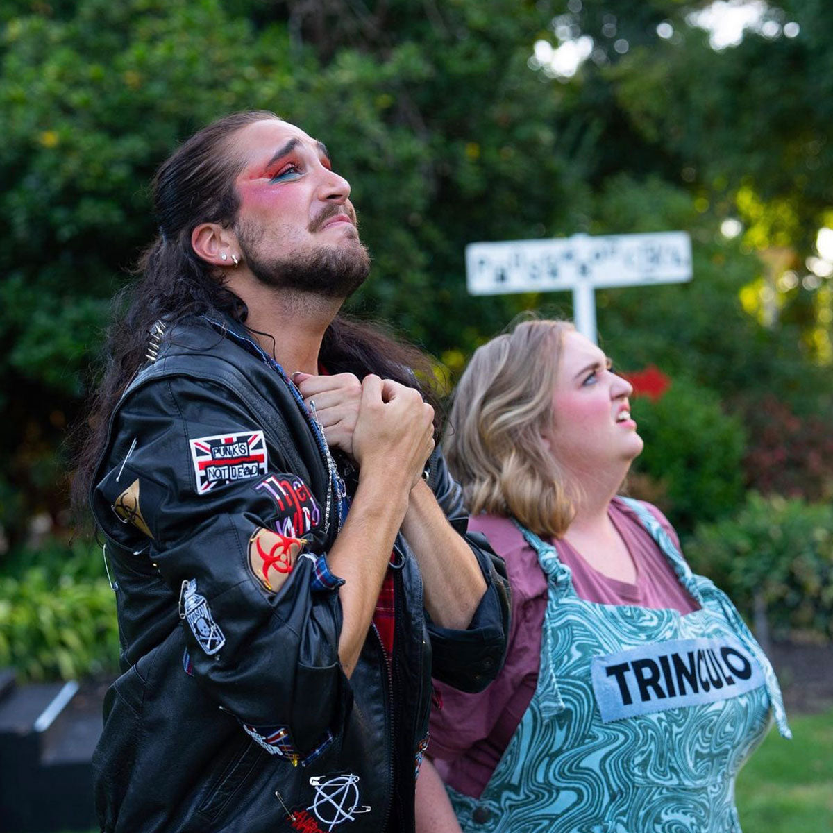 Mikey Sakinofsky - The Tempest_Mikey wearing leather jacket with patches on it, arms at chest with wrists crossed, looking skyward. Fellow actor beside them with apron on top of plum coloured t-shirt also looking skyward. Garden setting and trees in background.